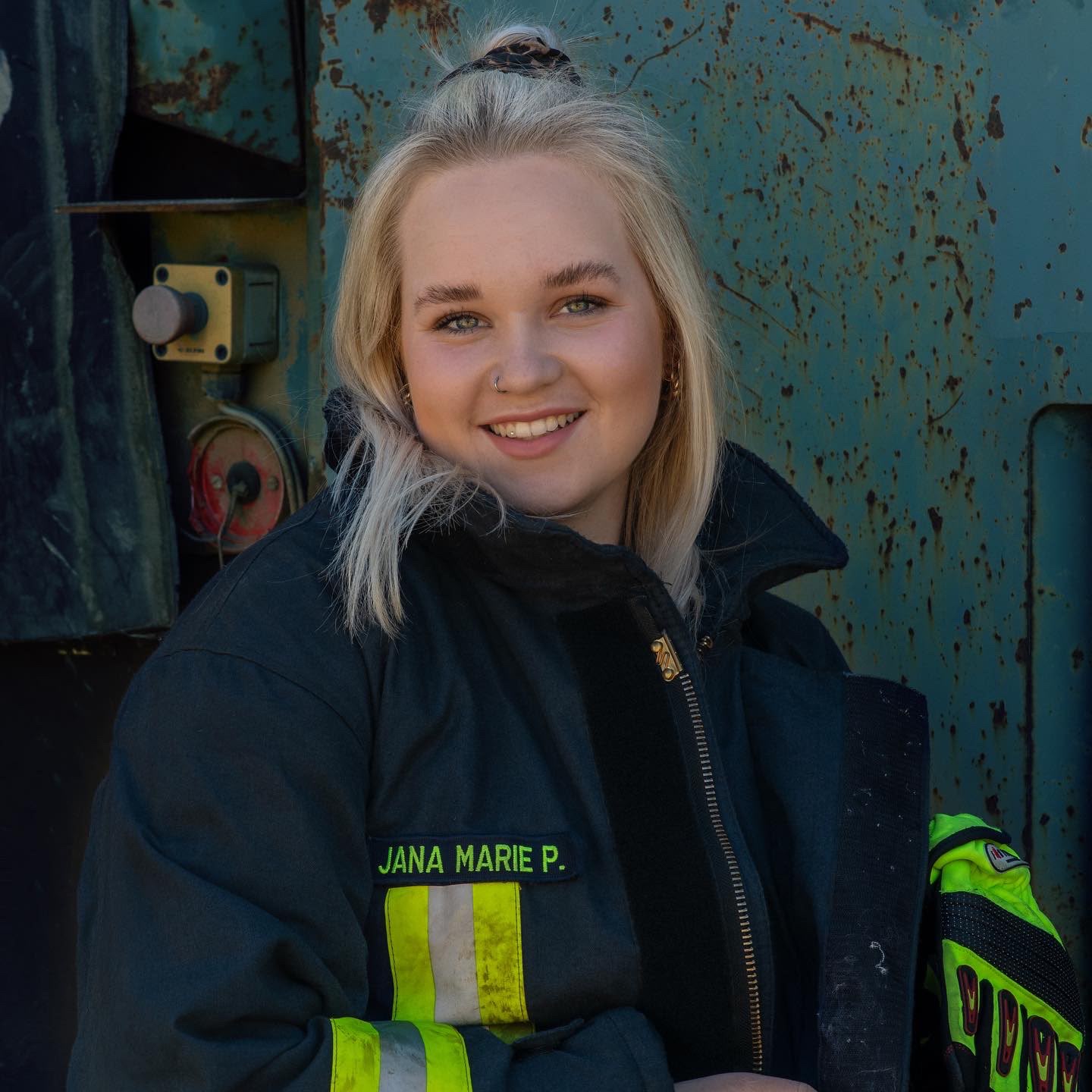 Jana Marie Paulsen in schwarzer Feuerwehrjacke mit reflektierenden Streifen, sitzend und lächelnd vor rostiger Metallwand.